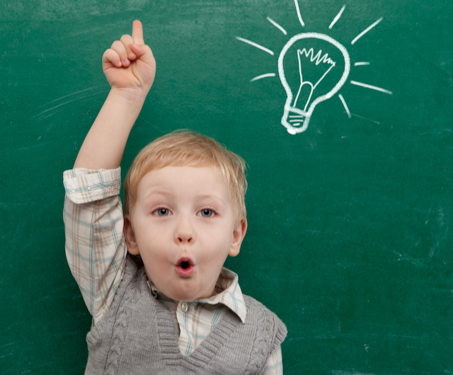 Young boy standing in front of green chalkboard, arm raised and finger pointing up. He appears to be saying, Oh! as if he has a great idea. 