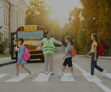 School crossing guard smiling and guiding elementary students across a crosswalk in front of a school bus.