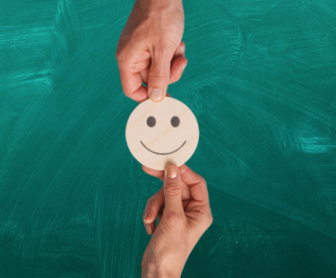 Smiley face exchanged between two hands, with green chalkboard behind it. 
