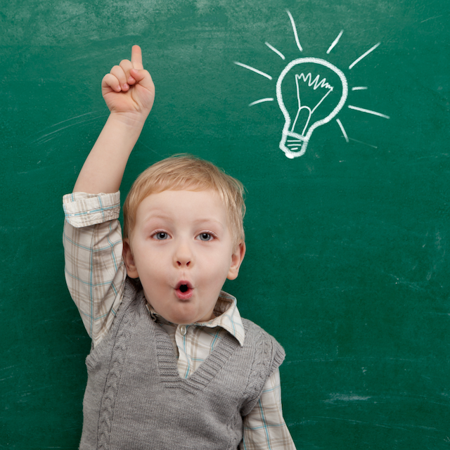 Young boy standing in front of green chalkboard, arm raised and finger pointing up. He appears to be saying, Oh! as if he has a great idea. 
