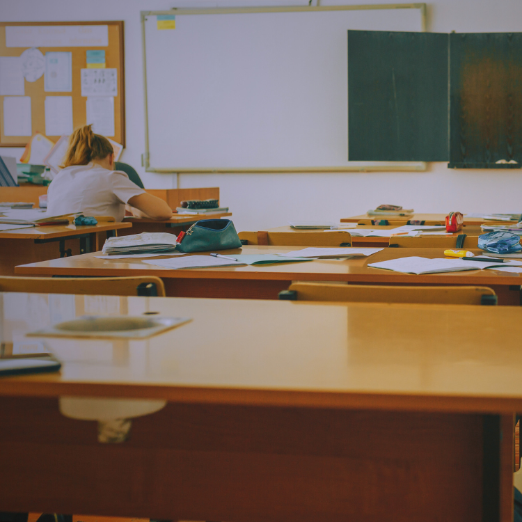 A nearly empty classroom with scattered papers and supplies on desks. A single teacher sits at the back, head down and working, suggesting fatigue or deep focus.
