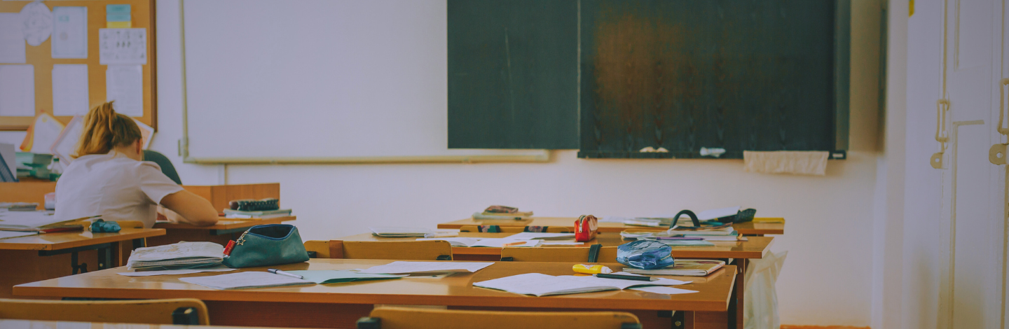 A nearly empty classroom with scattered papers and supplies on desks. A single teacher sits at the back, head down and working, suggesting fatigue or deep focus.