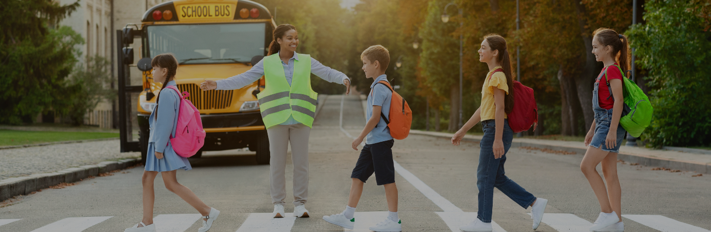School crossing guard smiling and guiding elementary students across a crosswalk in front of a school bus.