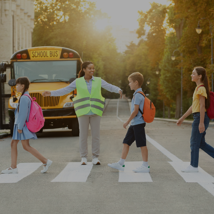 School crossing guard smiling and guiding elementary students across a crosswalk in front of a school bus.