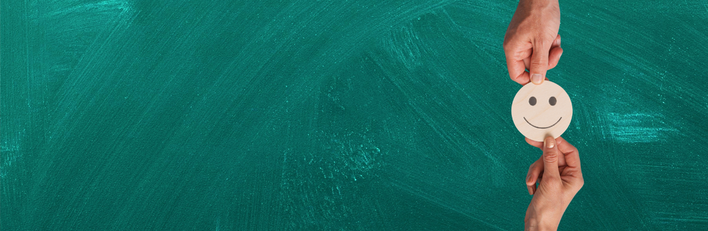 Smiley face exchanged between two hands, with green chalkboard behind it. 