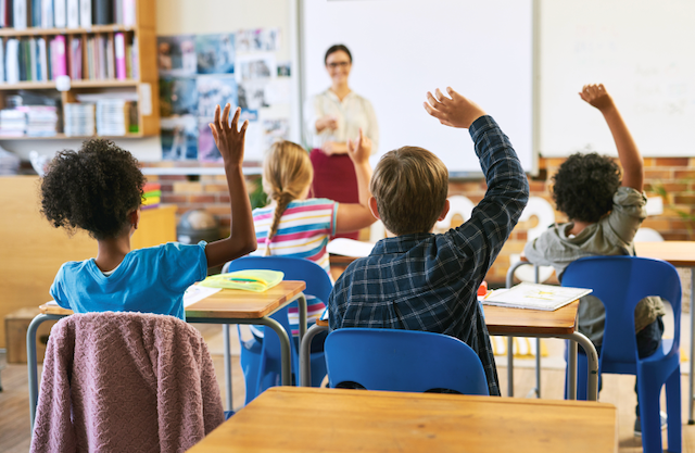 traditional classroom set-up. Teacher at front of classroom, students facing her, raising their hands.