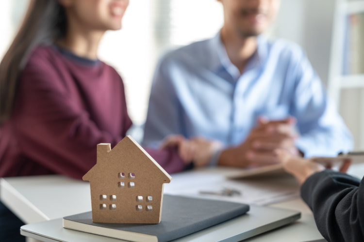 In foreground, a small, basic wooden model of a house. In blurred background, two parents looking across a desk at someone.